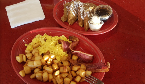 A red plate with scrambled eggs, diced potatoes, bacon, and a side of French toast with syrup and whipped cream.