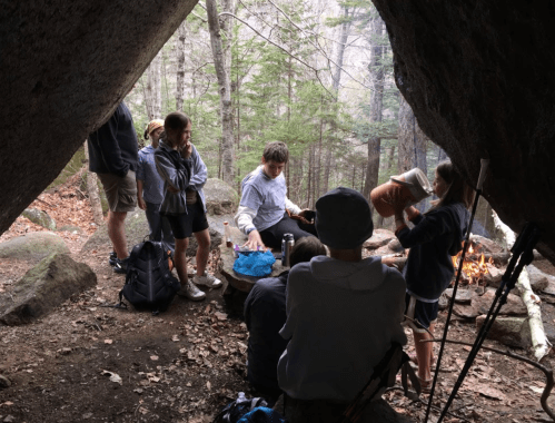 A group of people gathered around a campfire under a rock overhang in a forested area.