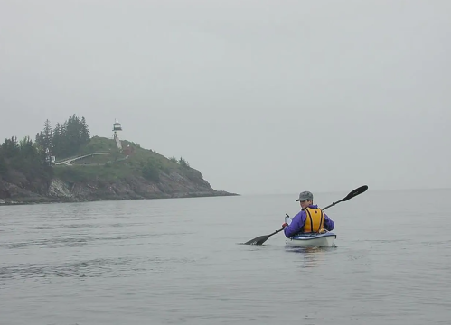 A kayaker paddles on calm waters near a foggy coastline with a lighthouse in the background.