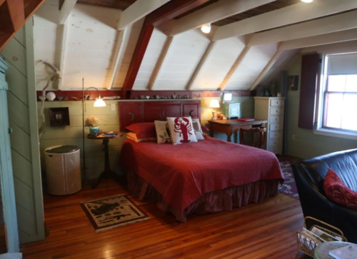 Cozy bedroom with a red bedspread, wooden beams, and a desk, featuring warm lighting and rustic decor.