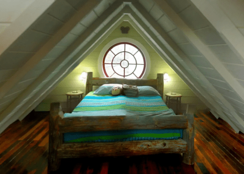 Cozy attic bedroom with a wooden bed, colorful bedding, and a round window, featuring sloped ceilings and wooden floors.