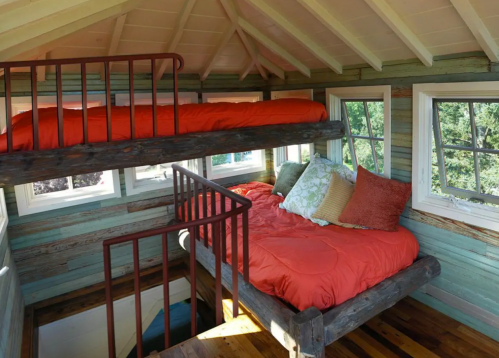 Cozy treehouse interior with two beds, wooden beams, and colorful cushions, surrounded by large windows.
