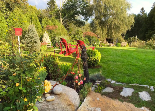 A serene garden scene featuring red chairs, lush greenery, and a stone pathway with flowers and a decorative pump.