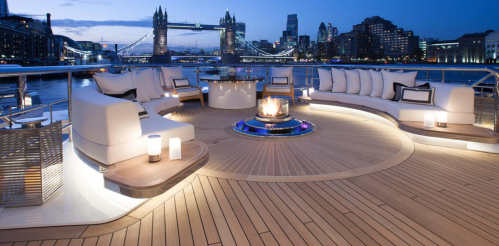 Luxurious yacht deck with modern seating, a fire pit, and city skyline views at dusk, featuring Tower Bridge in the background.