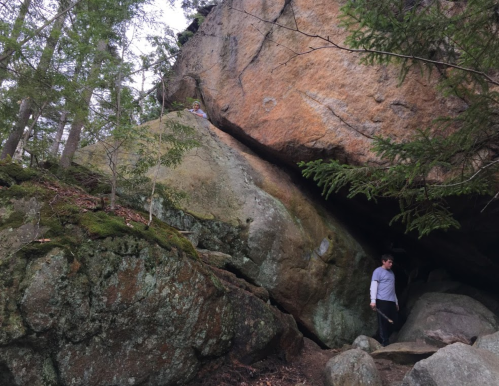 A person walking near a large rock formation in a forested area, with another person visible on a higher rock.