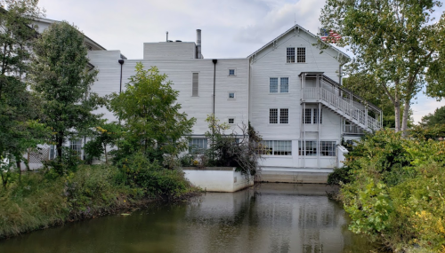 A white multi-story building beside a calm waterway, surrounded by trees and greenery, with an American flag visible.