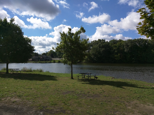 A serene lakeside scene with picnic tables, trees, and fluffy clouds in a blue sky.