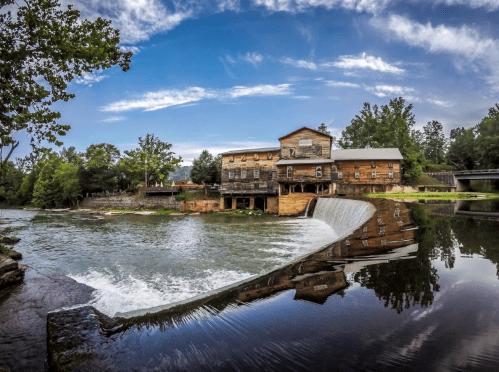A historic wooden mill beside a flowing river, surrounded by lush greenery and a clear blue sky.