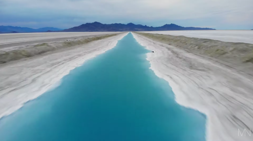Aerial view of a vibrant turquoise waterway bordered by white salt flats and distant mountains under a cloudy sky.