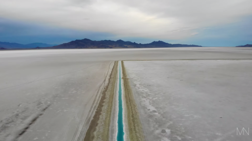 Aerial view of a vast salt flat with a narrow blue waterway cutting through the dry landscape and distant mountains.