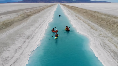Aerial view of kayakers paddling through a bright blue canal surrounded by white salt flats.