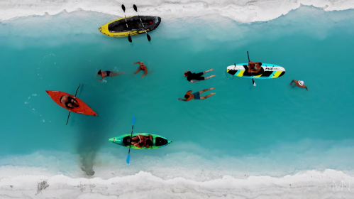 Aerial view of people kayaking and swimming in a turquoise lagoon surrounded by white sand.