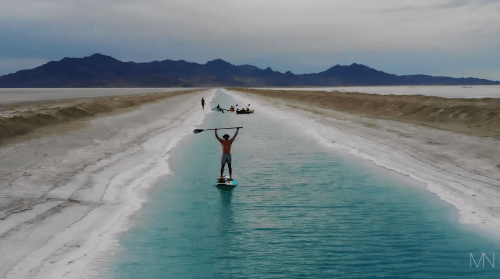 A person paddleboarding in a narrow, turquoise waterway with mountains in the background and others kayaking nearby.
