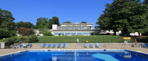 A clear blue pool in the foreground with lounge chairs, set against a modern building and green trees under a blue sky.