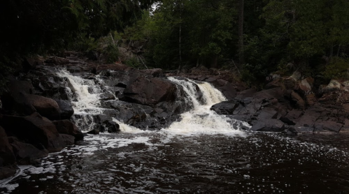 A serene waterfall cascades over dark rocks, surrounded by lush green trees and a calm, dark water pool below.