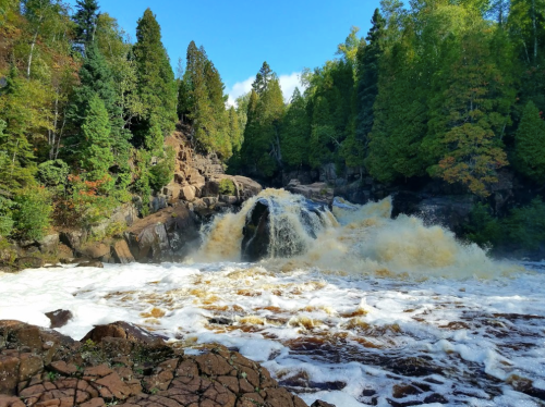 A cascading waterfall surrounded by lush green trees and rocky terrain under a clear blue sky.
