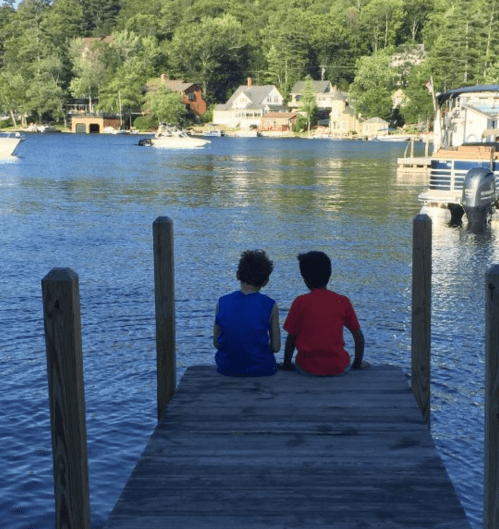 Two children sit on a wooden dock, looking out over a calm lake with houses and boats in the background.