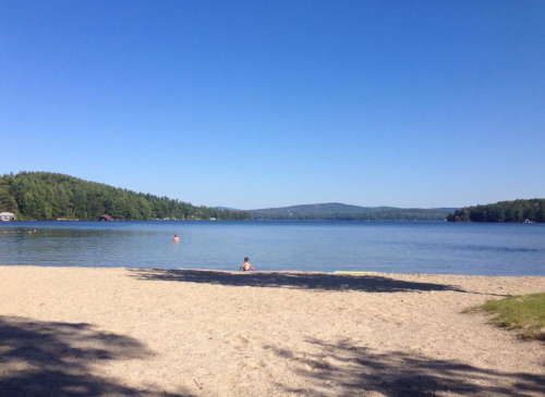 A serene lake scene with a sandy beach, clear blue water, and green hills under a bright blue sky.