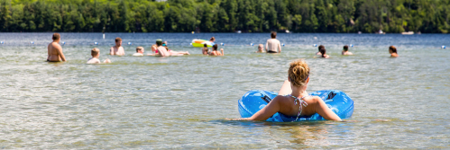 A woman floats on a blue inner tube in a lake, surrounded by people swimming and enjoying the water on a sunny day.