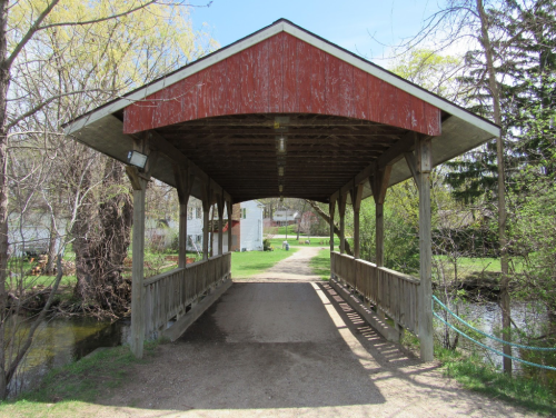 A wooden covered bridge leads to a grassy area with trees and a white building in the background.