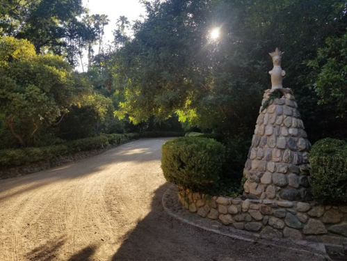 A winding dirt path surrounded by lush greenery, featuring a stone sculpture at the edge of the road.