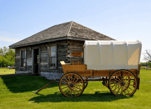 A wooden wagon with a white cover parked beside a rustic log cabin on a grassy field under a clear blue sky.