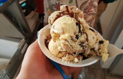 A hand holding a bowl of ice cream with various flavors and chocolate syrup on top.