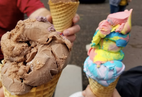 Two ice cream cones held by hands: one with chocolate and the other with colorful rainbow flavors.
