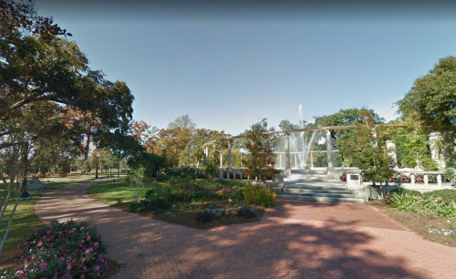 A scenic garden with a fountain, surrounded by colorful flowers and trees, under a clear blue sky.