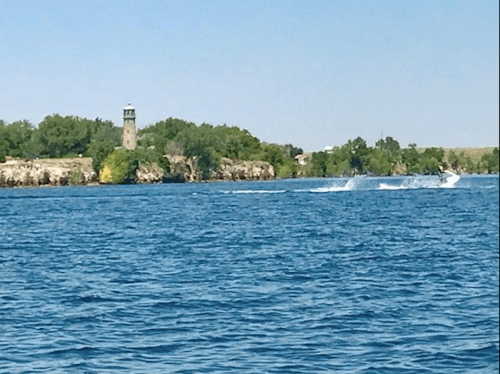 A scenic view of a lake with a distant tower and a person water skiing, surrounded by greenery and clear blue skies.