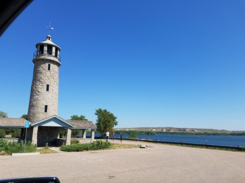 A stone lighthouse beside a calm lake, with clear blue skies and a few people walking nearby.