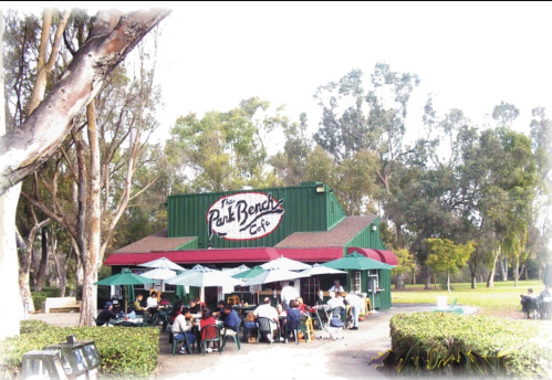 A café with outdoor seating under umbrellas, surrounded by trees and greenery, bustling with people enjoying their meals.