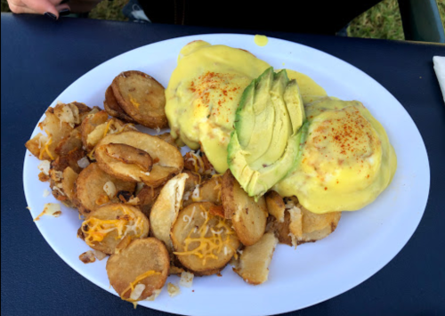 A plate of eggs Benedict topped with avocado and hollandaise sauce, served with crispy potato slices.