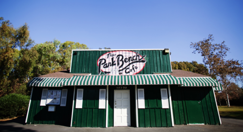 A green building with a striped awning, featuring a sign that reads "The Park Bench Café" against a clear blue sky.