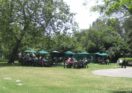A sunny park scene with people sitting at tables under green umbrellas, surrounded by trees and grass.
