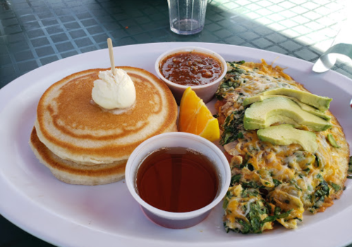 A plate with pancakes topped with butter, an omelet with avocado, salsa, orange slice, and syrup on the side.