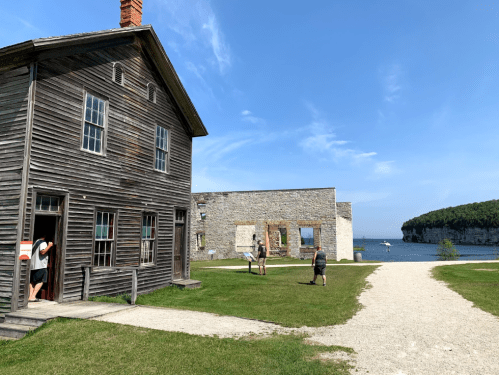 A wooden building and stone ruins by the water, with people walking on a grassy path under a clear blue sky.
