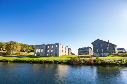 Historic buildings beside a calm river, with clear blue skies and green grass in the foreground.