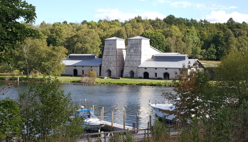 Historic stone buildings by a calm river, surrounded by lush greenery and boats docked nearby.