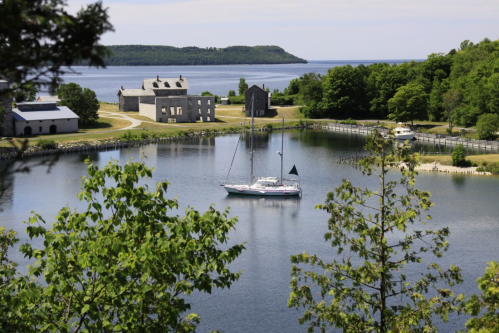 A serene harbor scene with a sailboat, historic buildings, and lush greenery under a clear sky.