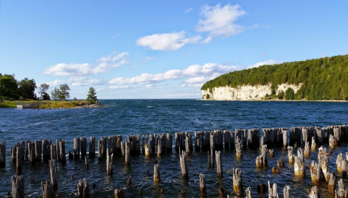 A scenic view of a lake with wooden posts in the foreground and a lush, rocky shoreline under a blue sky.