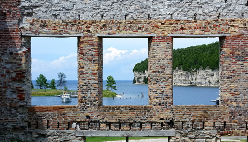 View through brick ruins to a serene lake with a boat and lush green cliffs in the background.