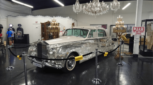 A vintage car covered in mirrors, displayed in a museum with chandeliers and a "Do Not Touch" sign nearby.