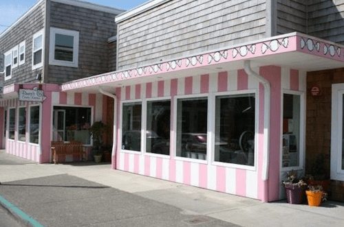 A pink and white striped storefront with large windows and potted plants outside, featuring a wooden bench.