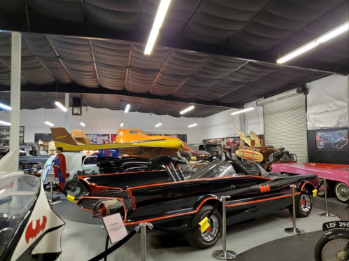 A vintage car showroom featuring a black Batmobile and various classic cars and planes on display.