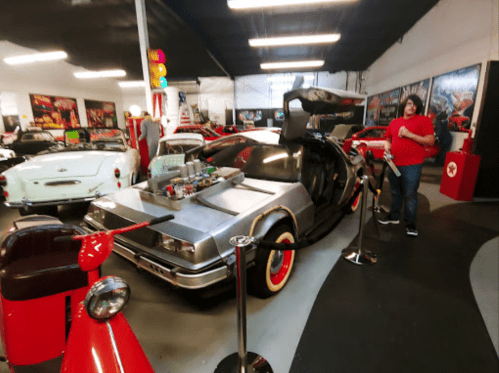 A vintage car exhibit featuring a DeLorean with gullwing doors, surrounded by classic cars and visitors.