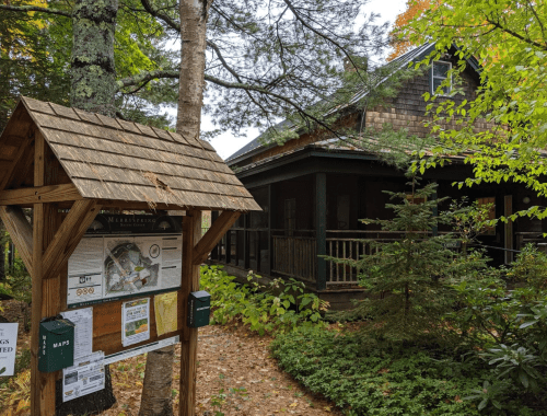 A wooden sign with a map stands in front of a rustic cabin surrounded by trees and autumn foliage.