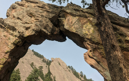 Natural rock arch framed by trees, with rugged mountains in the background under a clear sky.