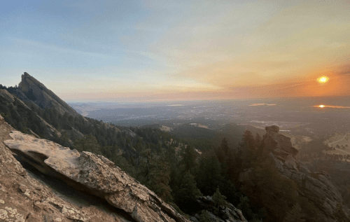 Sunset view from a mountain peak, overlooking a valley with trees and distant hills, bathed in warm golden light.
