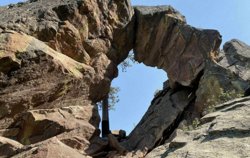 A natural rock arch framed by rugged stone, with a clear blue sky above and greenery peeking through the opening.